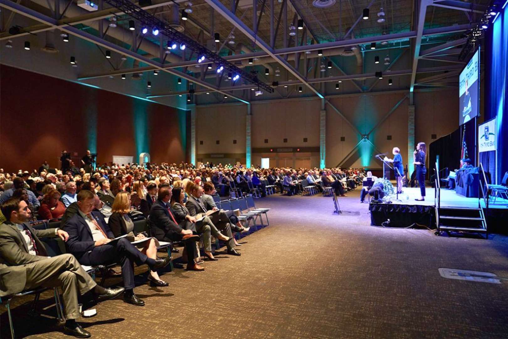 Photo of Audience sitting at a conference in One National Events Plaza as a woman addresses them on stage