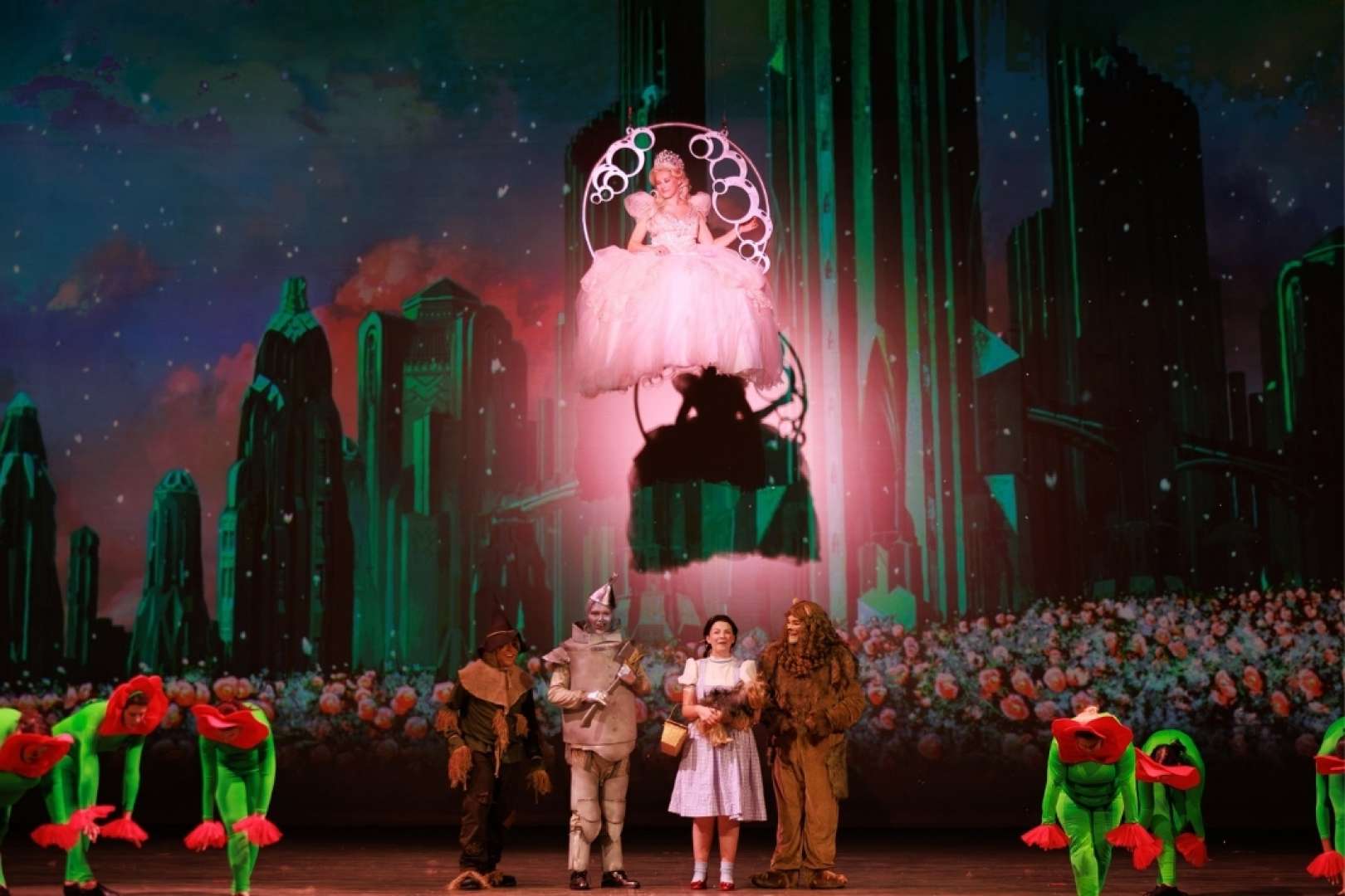 Photo of A vibrant stage production of The Wizard of Oz by the Paramount Players, featuring Dorothy, the Tin Man, the Cowardly Lion, the Scarecrow, and Toto, while Glinda the Good Witch watches over them from above.