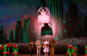 Thumbnail photo of A vibrant stage production of The Wizard of Oz by the Paramount Players, featuring Dorothy, the Tin Man, the Cowardly Lion, the Scarecrow, and Toto, while Glinda the Good Witch watches over them from above.