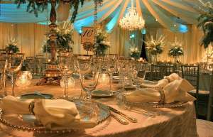 Thumbnail photo of A close-up captures a beautifully arranged wedding table, elegantly set for dinner. In the background, a grand chandelier and towering floral arrangements add a touch of sophistication and charm.